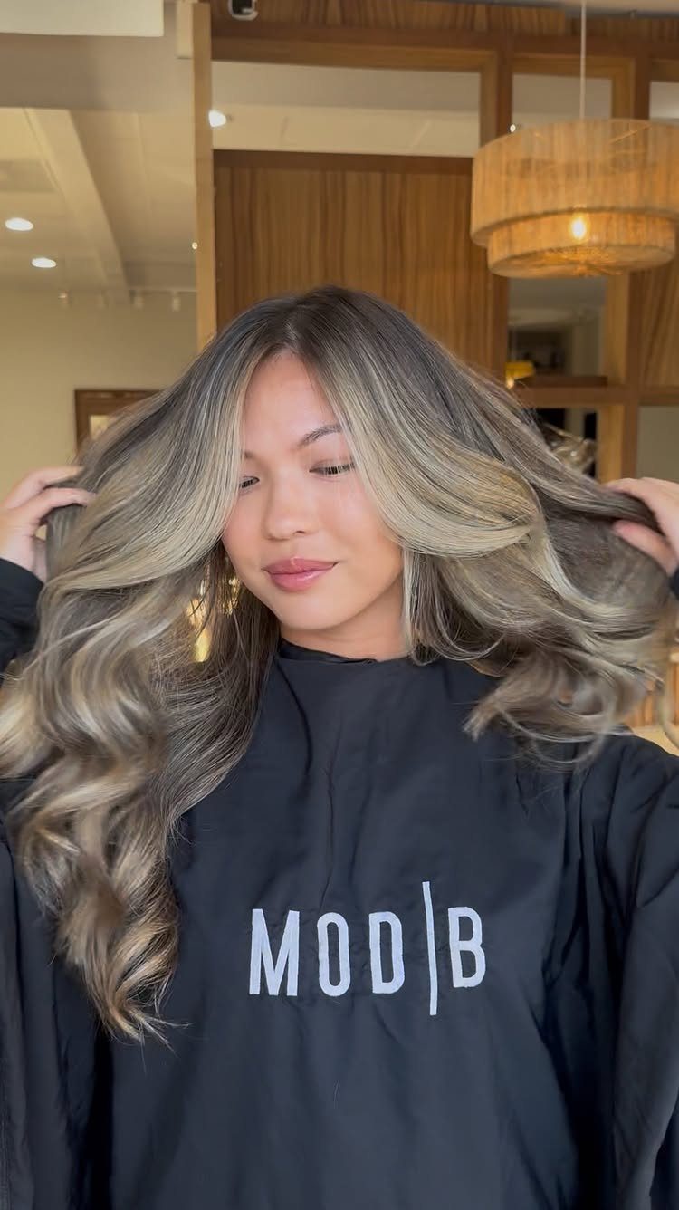 Woman displaying long ash-brown waves with darker melted roots in a salon