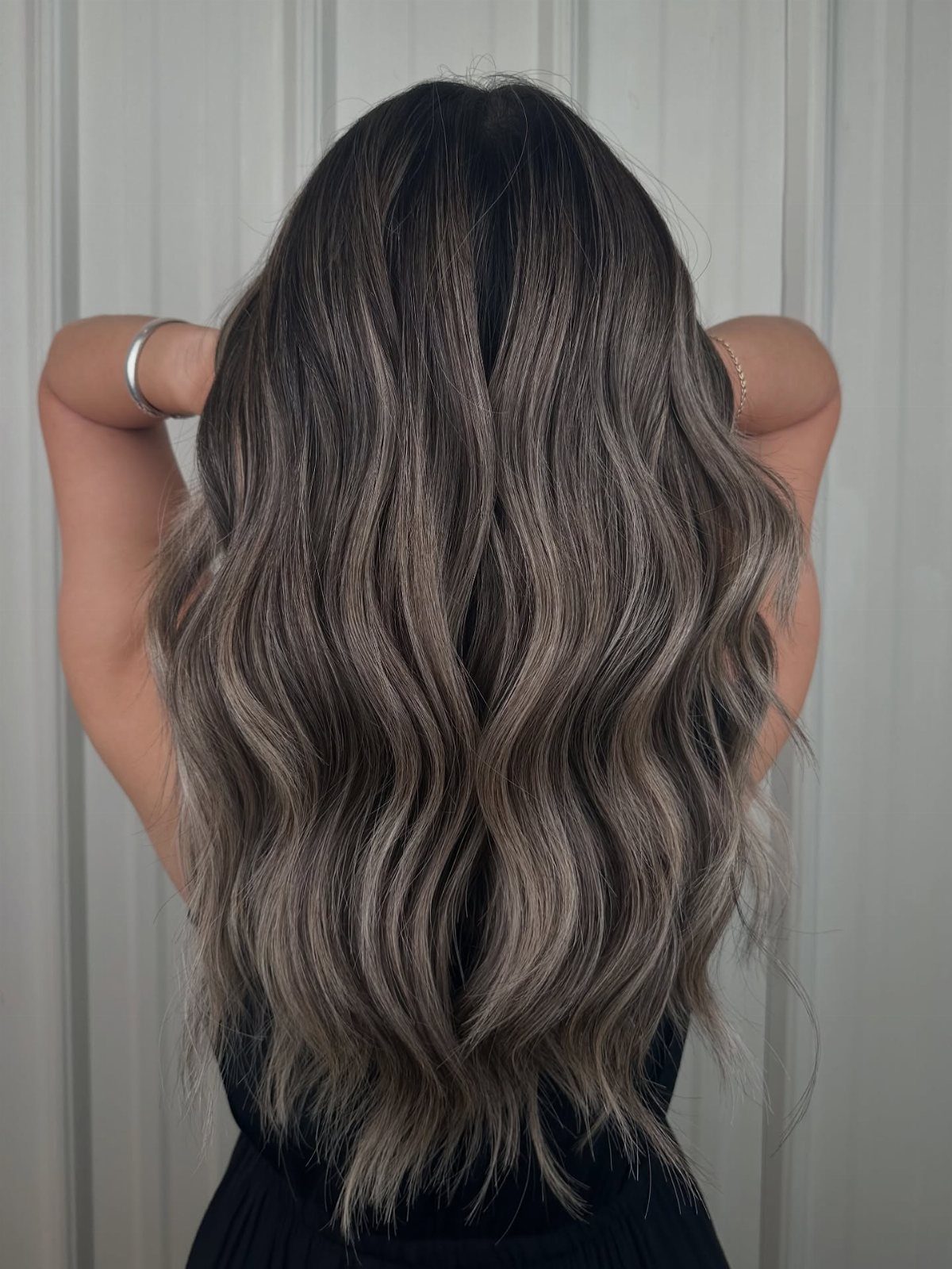Long brunette waves show cool metallic taupe balayage ribbons against a pale paneled wall