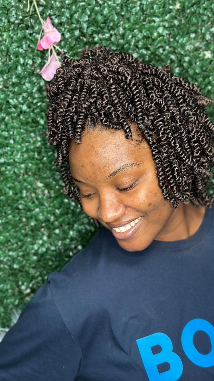Woman with short black crochet braids smiles while looking down against leafy greenery