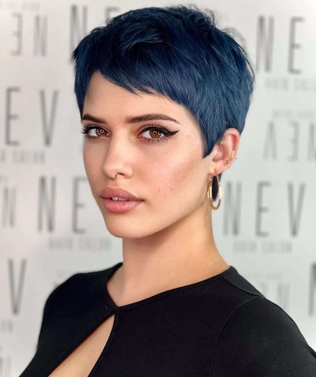 Woman posing with a short, close-cropped blue-black pixie haircut and hoop earrings