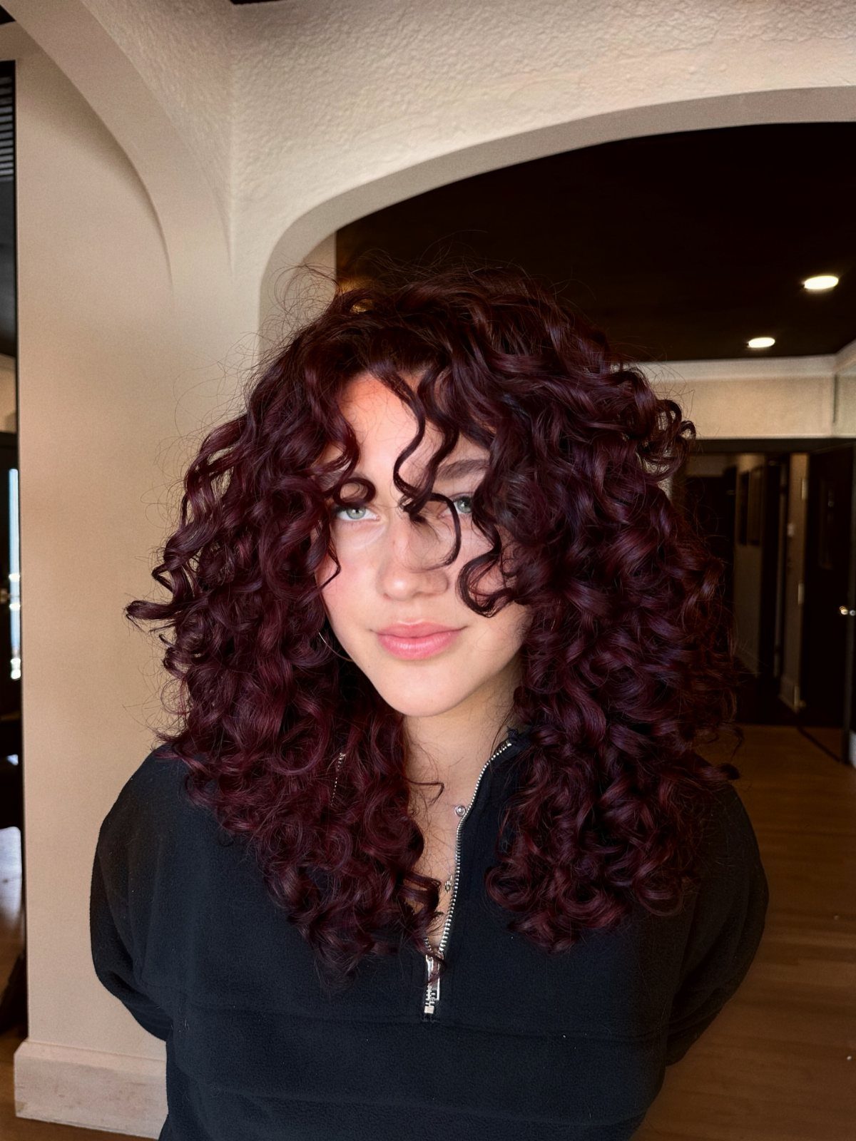 Person with glossy wine-red curls standing beneath a white indoor archway
