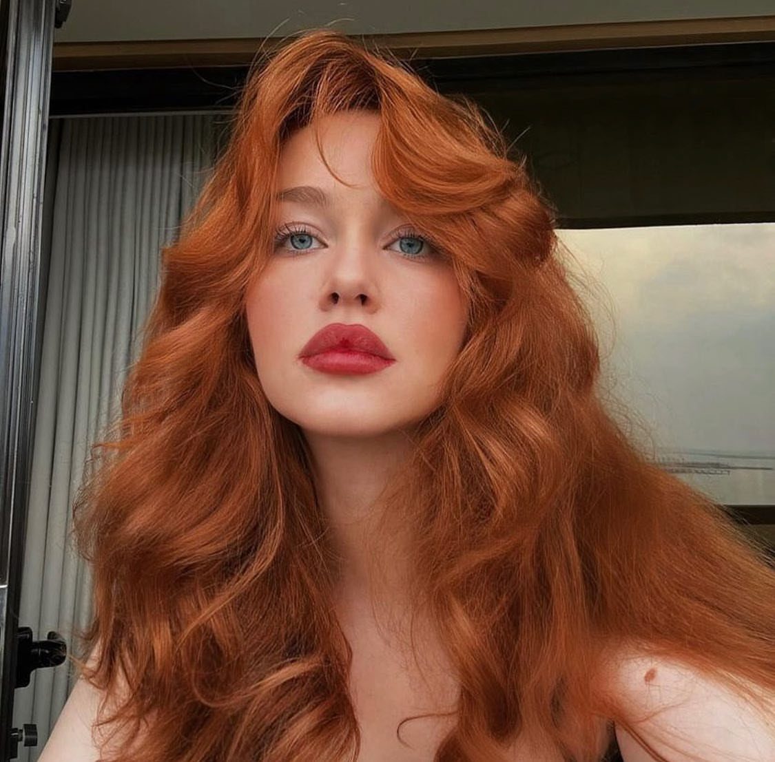 Woman with long, voluminous bright ginger-copper waves in front of a window