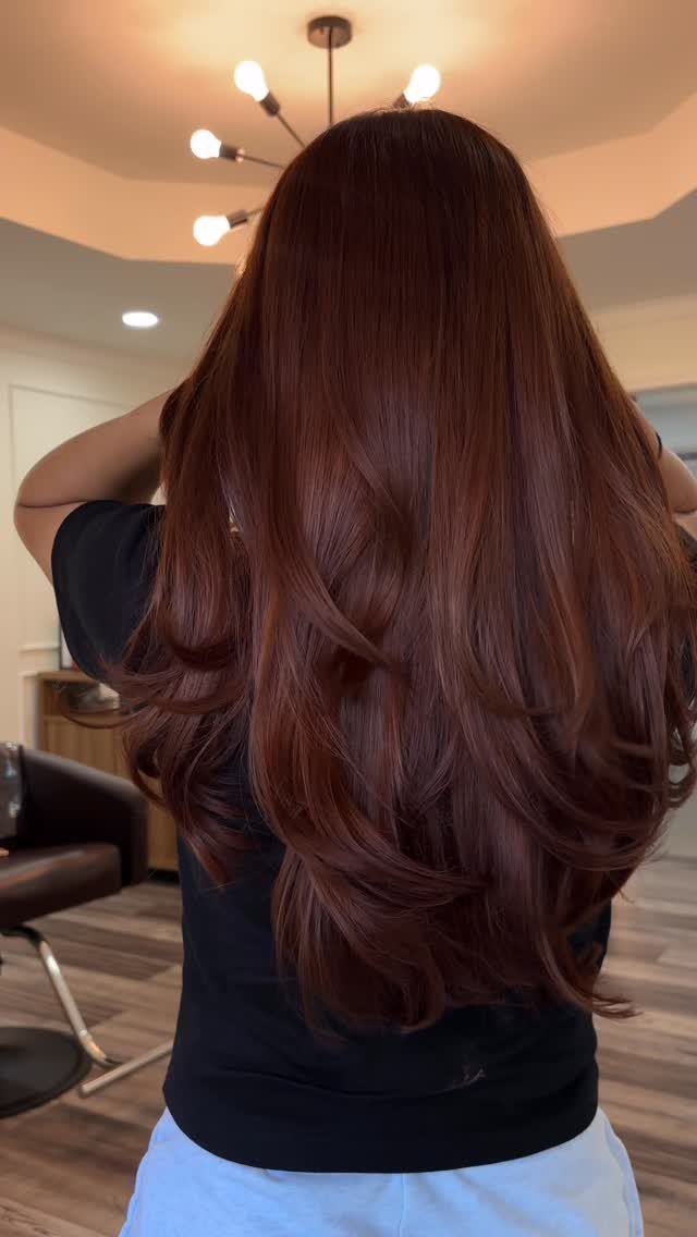 Long layered copper-brown hair shines under salon lights against a dark top.