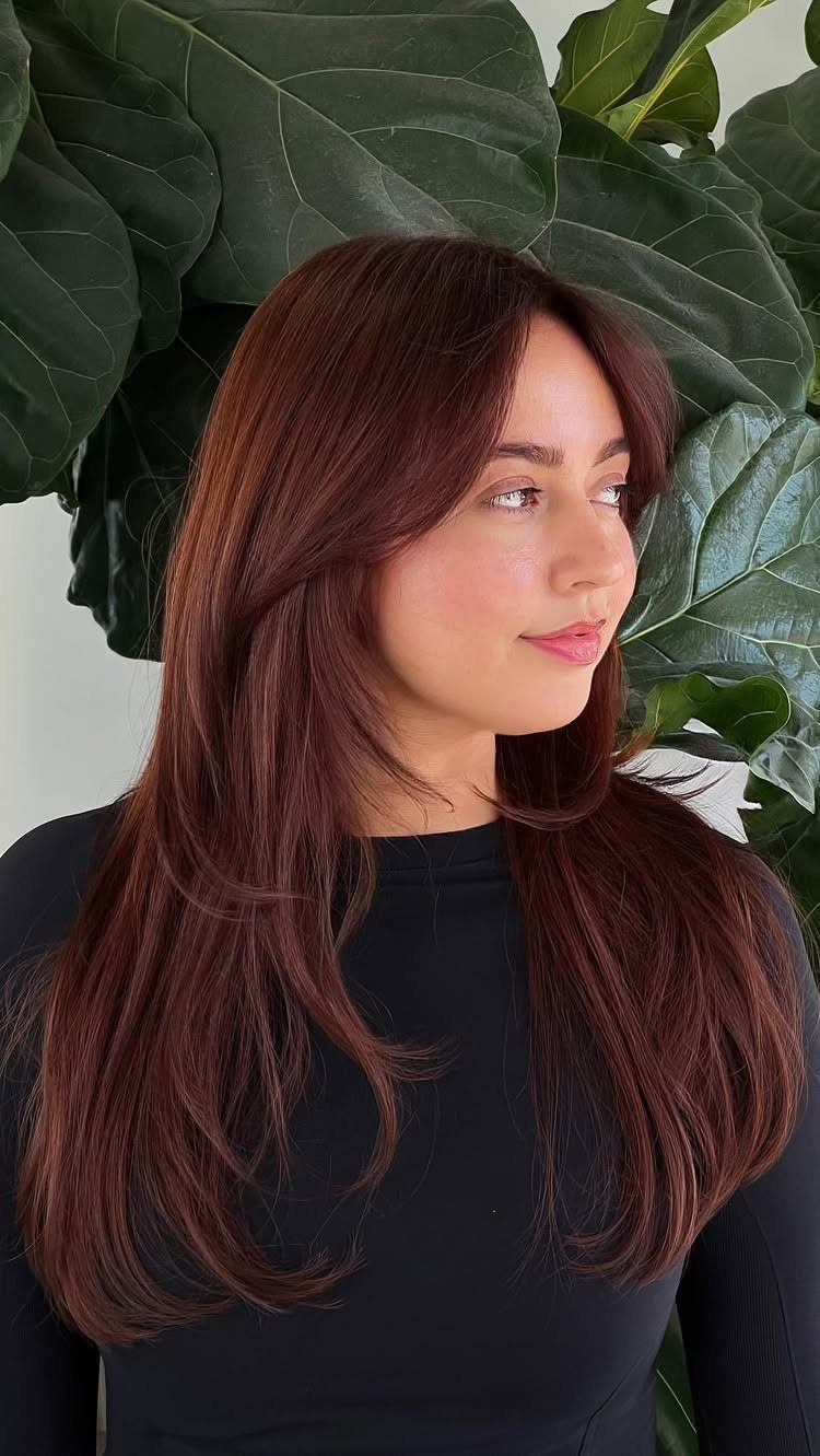 Woman with long brown copper-toned hair posing in front of leafy greenery