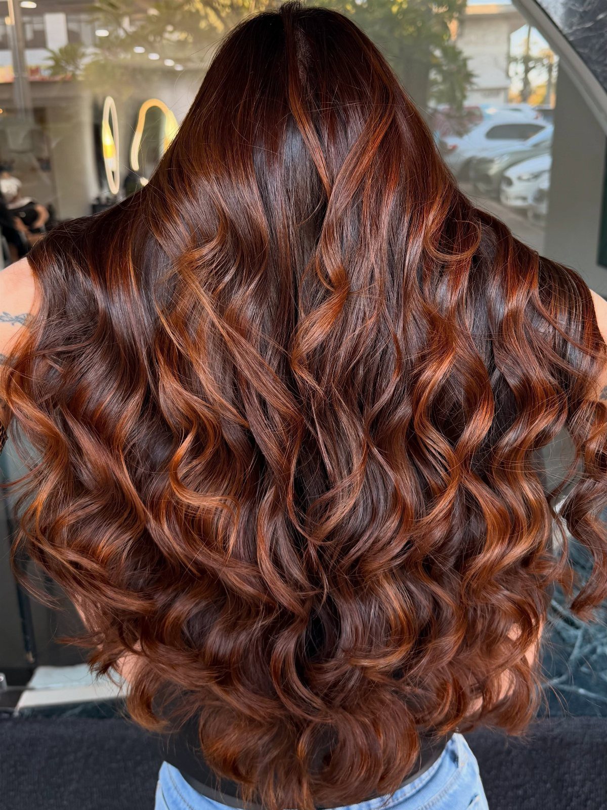 Long curled auburn-brown hair with warm copper highlights, photographed from behind in a salon