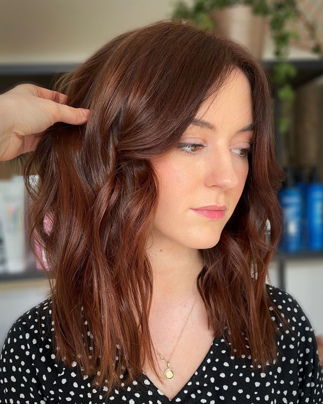Woman with softly waved reddish-brown hair in a salon, a stylist lifting one section