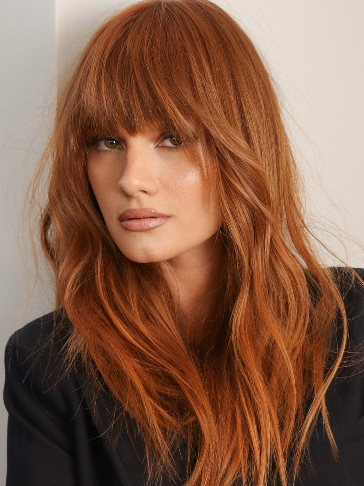 Woman with long copper-auburn layers and full bangs against a light wall