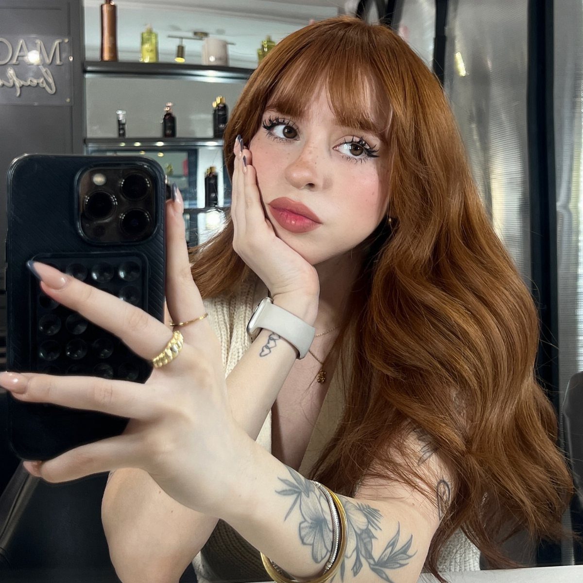Woman showing long golden copper-brown waves with bangs in a salon mirror selfie