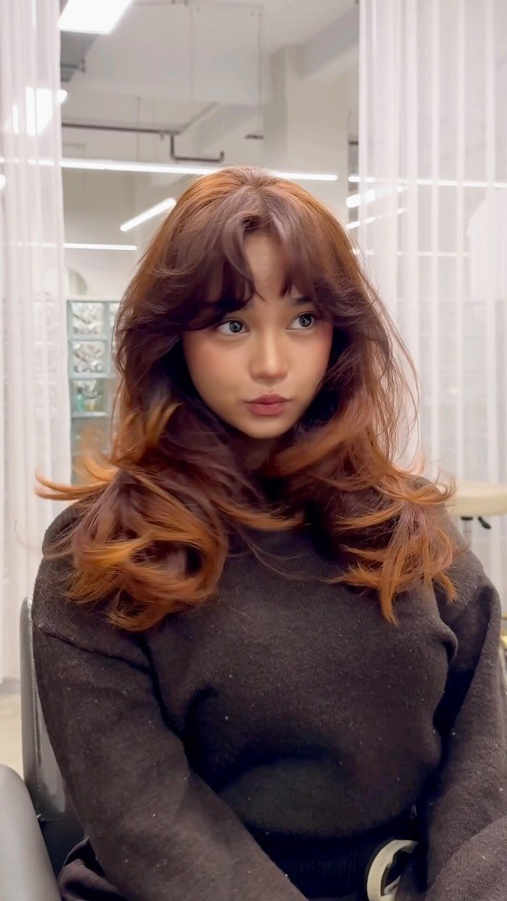 Woman displaying long copper-brown waves and curtain bangs in a bright salon