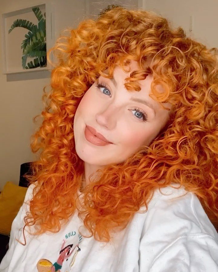 Person with voluminous copper-orange curls and bangs posing indoors