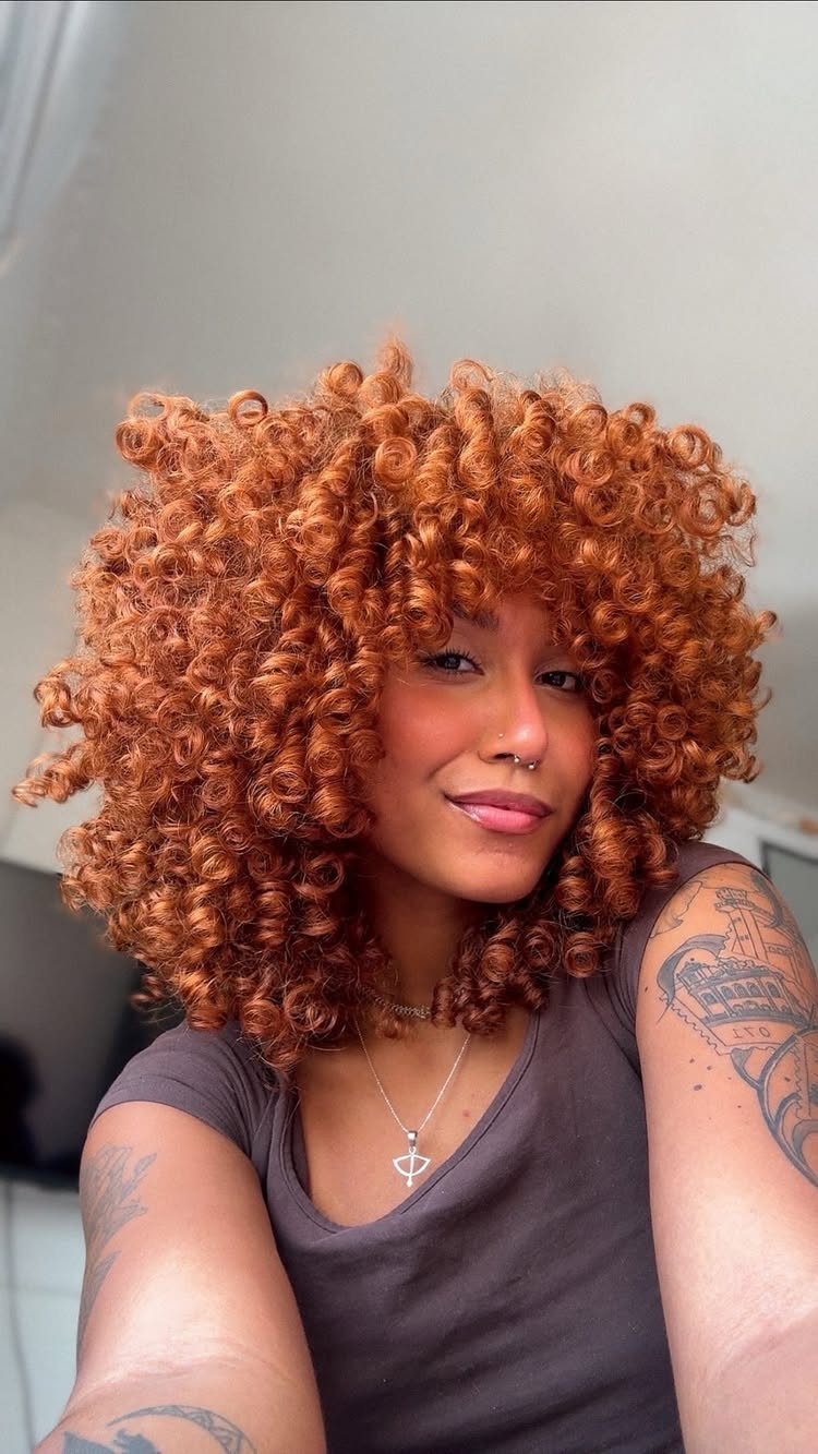 Woman with voluminous copper-orange curls poses indoors in a dark sleeveless top