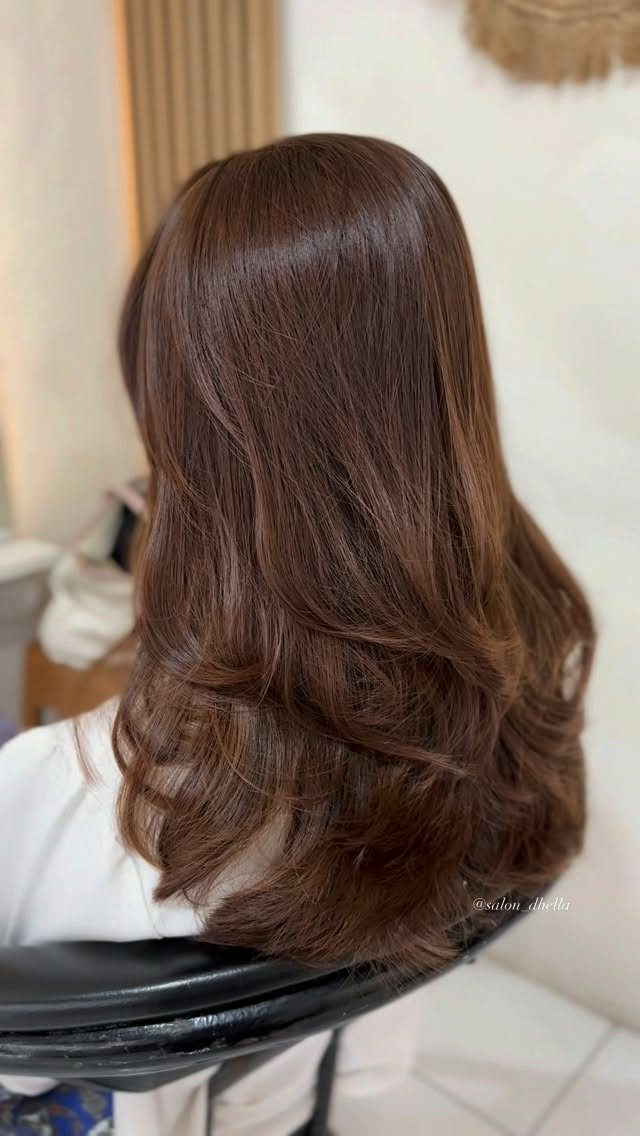 Long brunette hair falls in glossy, softly curled layers over a white salon cape.