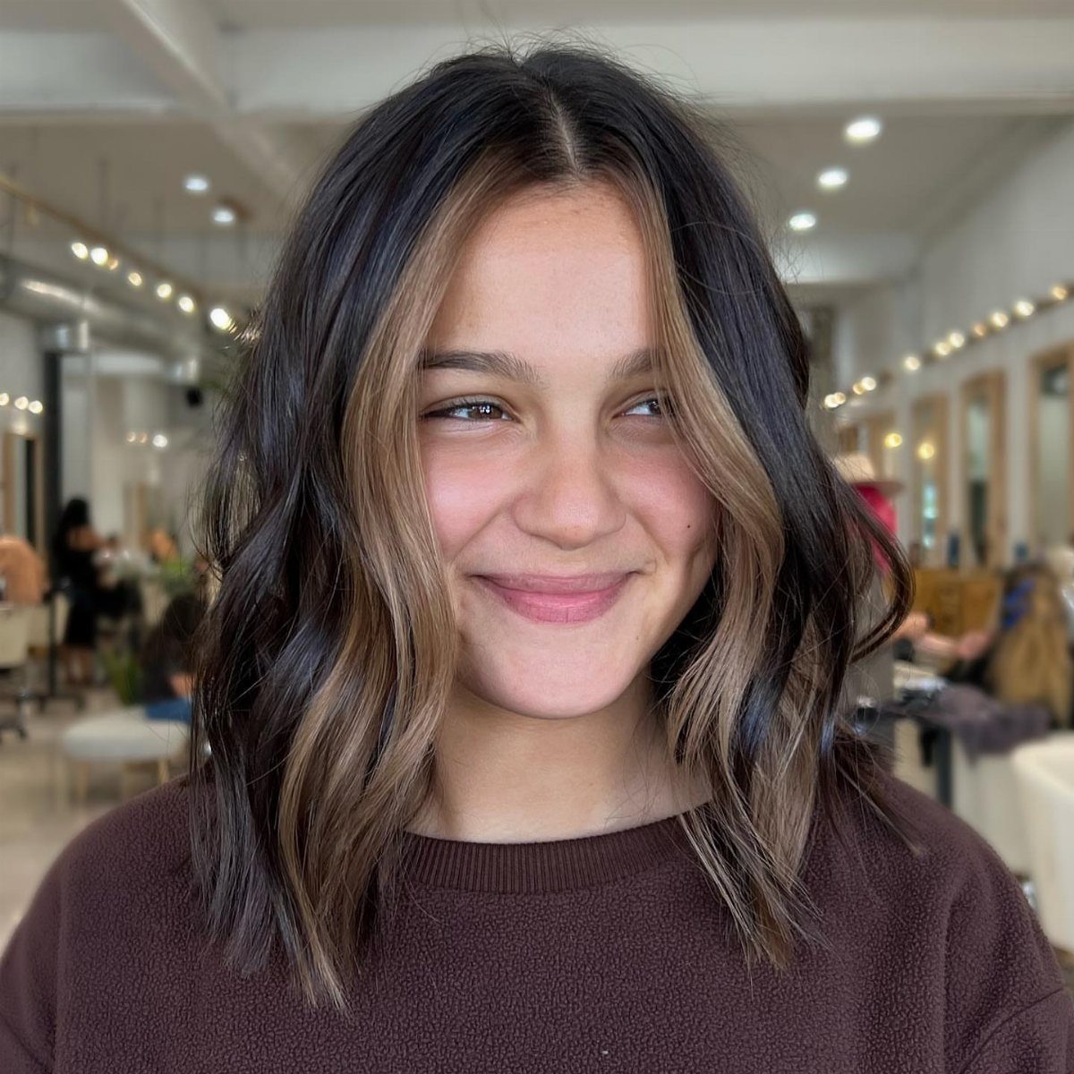 Woman with wavy dark brown hair and lighter face-framing highlights in a salon