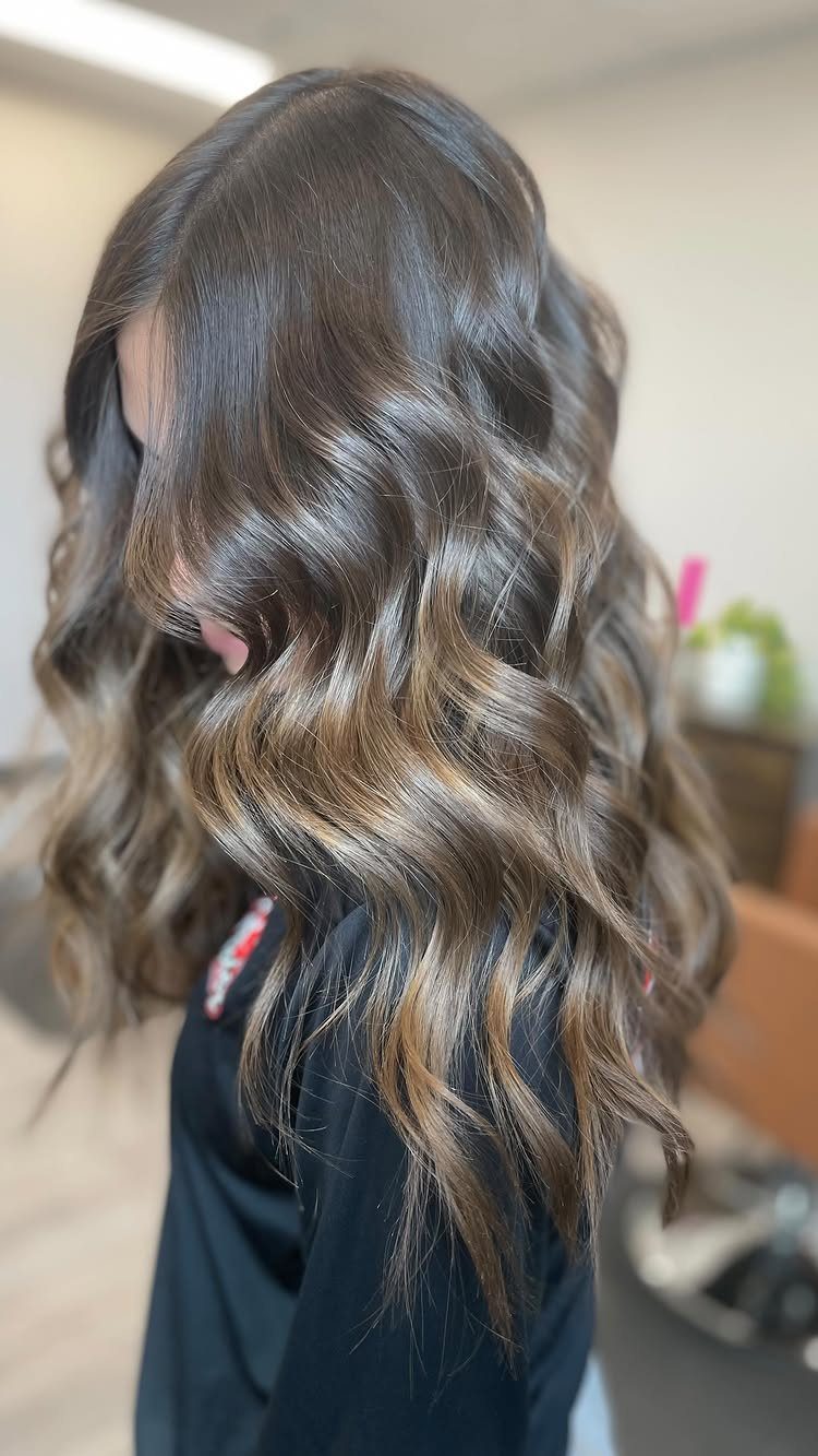 Wavy dark brown hair with warm golden-brown highlights cascading down a salon cape
