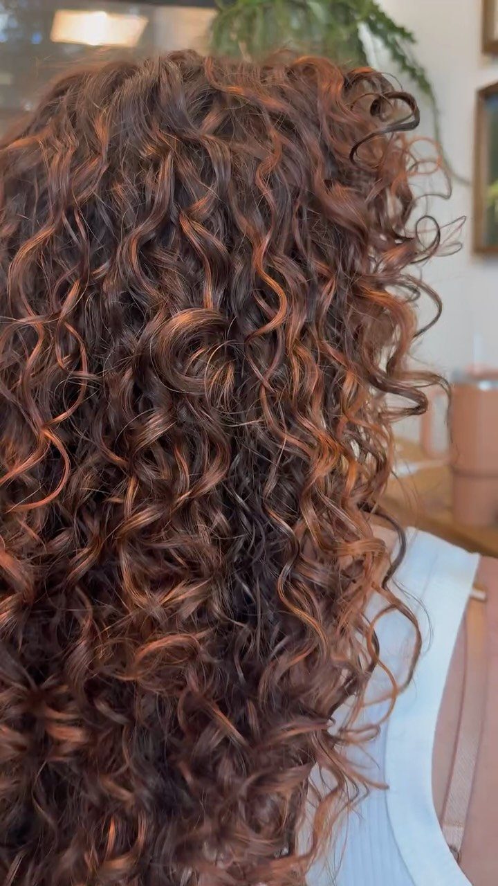 Brown curly hair with warm coral-red highlights cascading over a light blue top