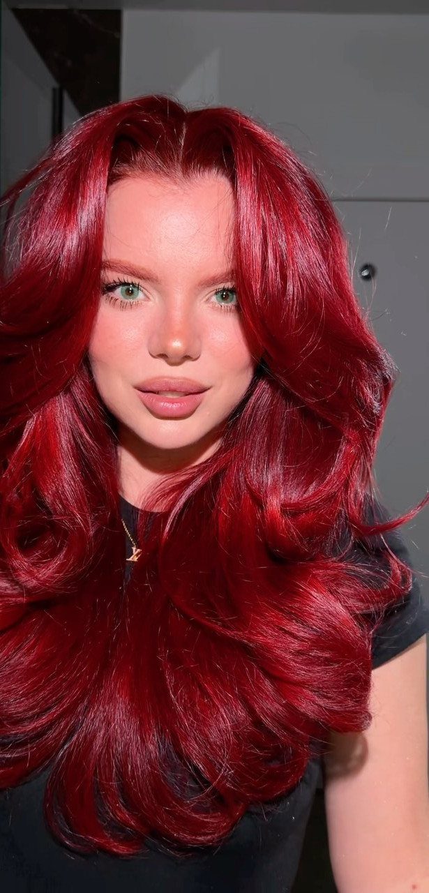 Person with long, glossy deep crimson hair posing against a gray wall