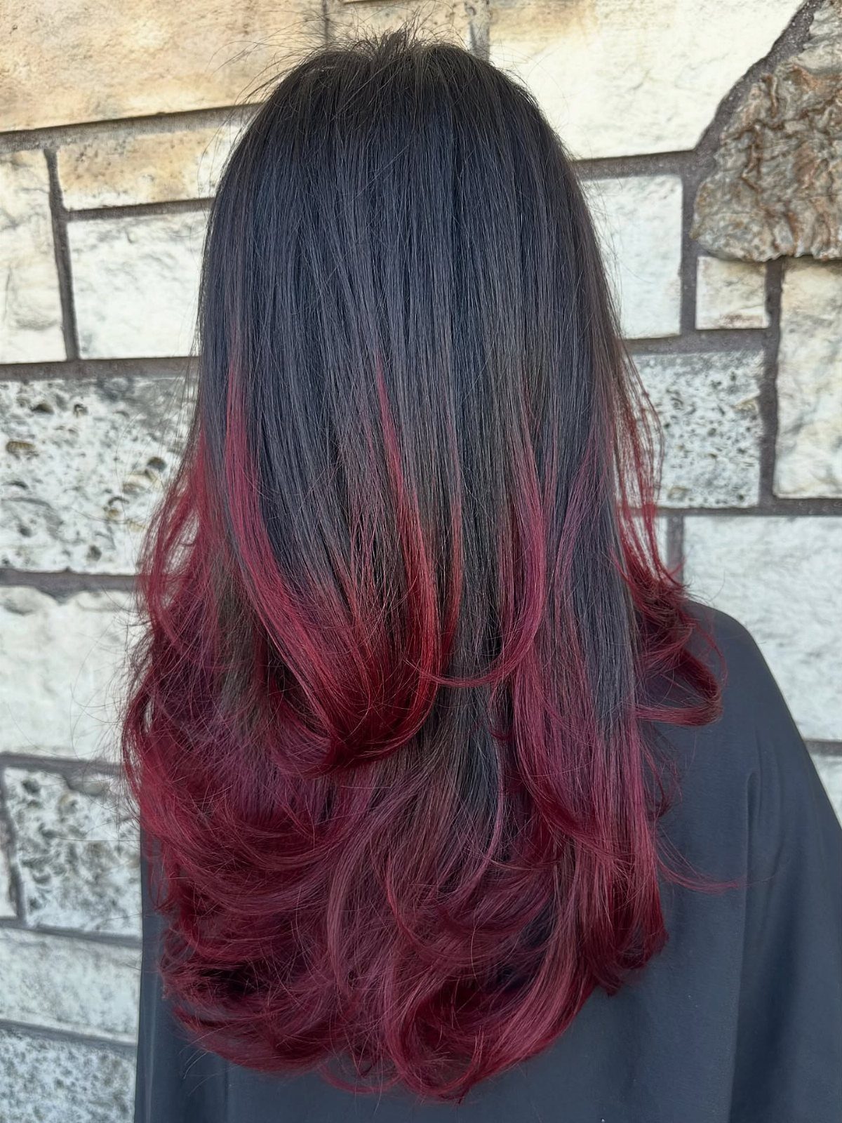 Long layered hair with dark roots and deep wine-red ends against a stone wall