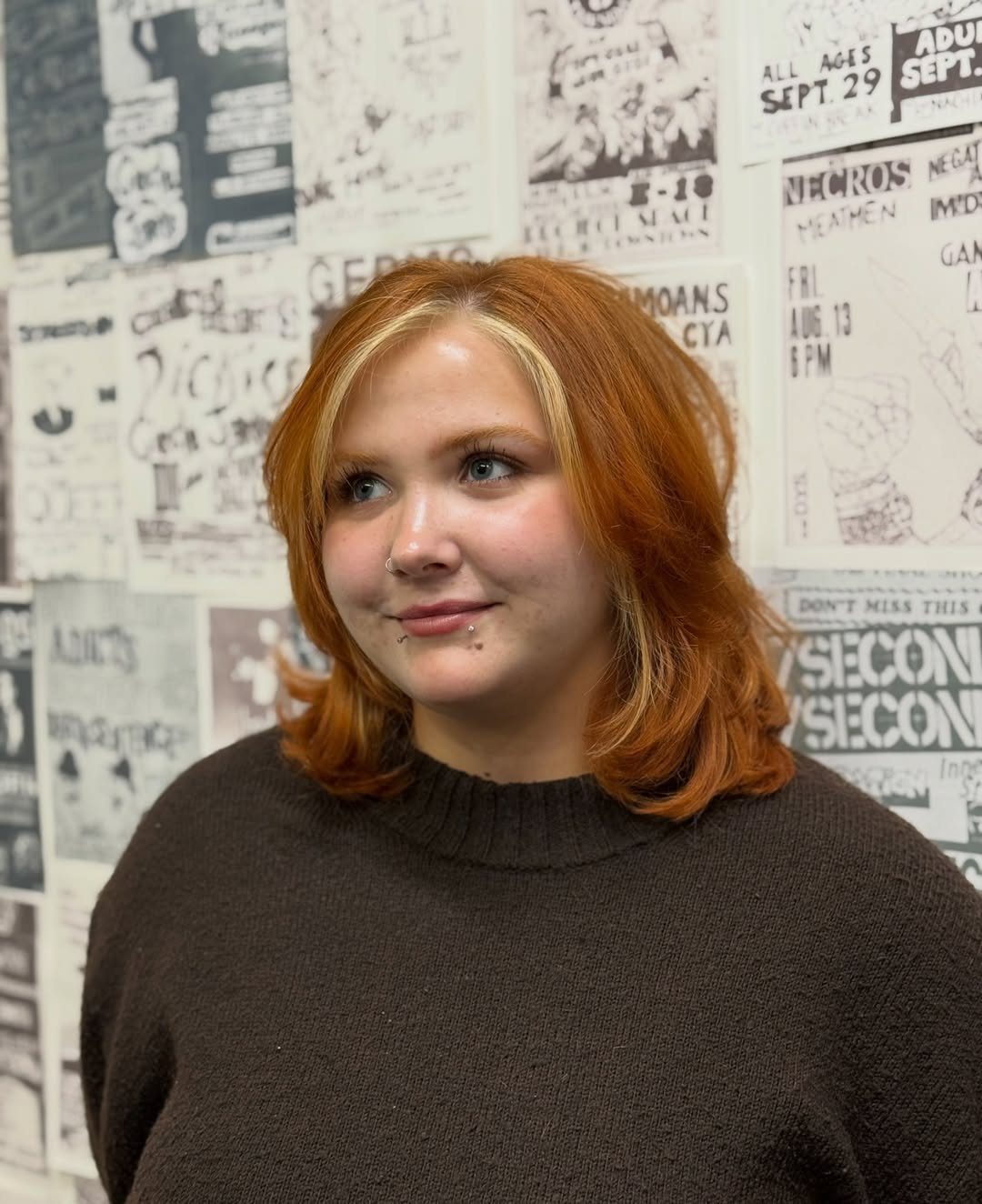 Person with copper-orange hair and lighter face-framing strands against a poster-covered wall