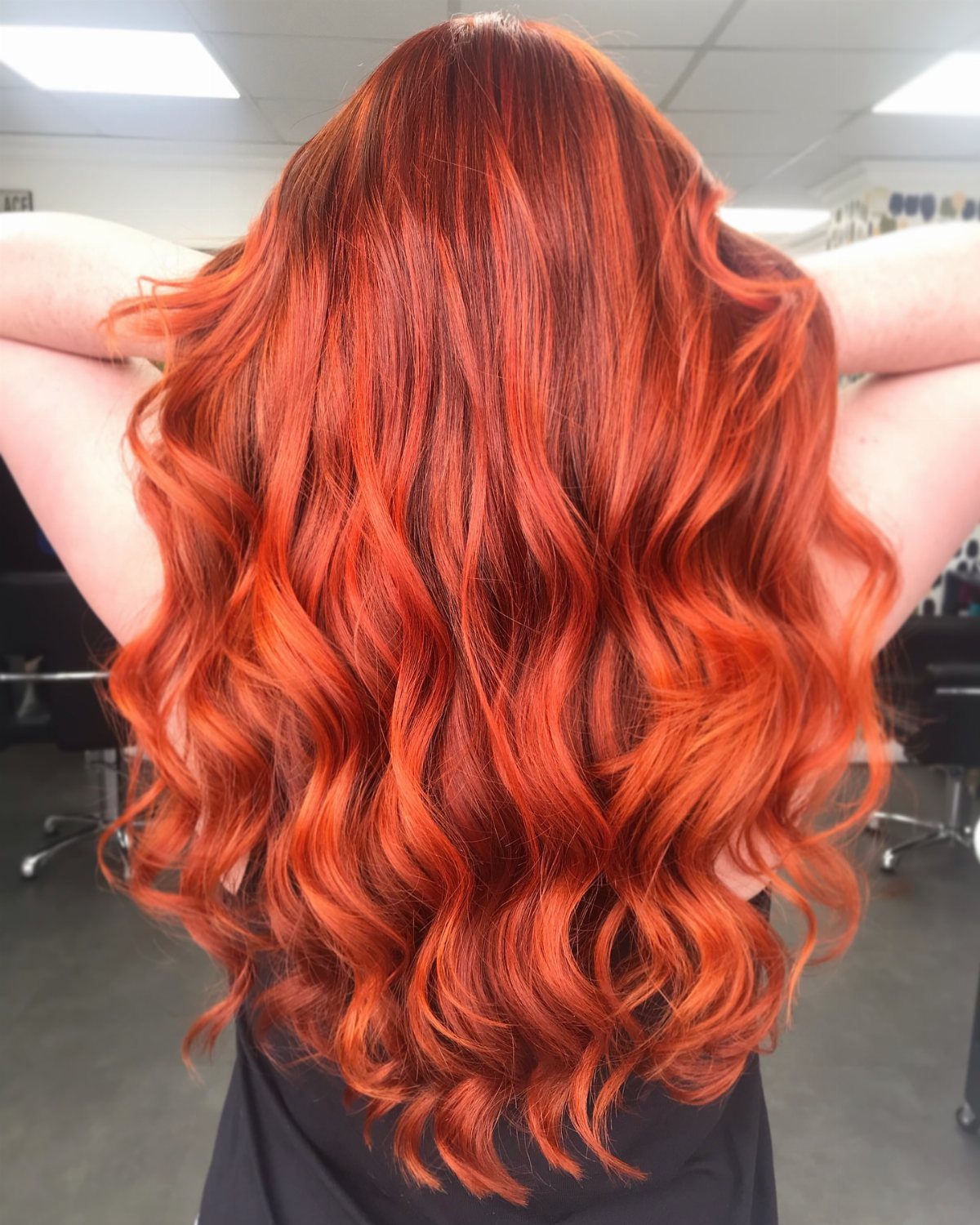Long wavy bright orange-red balayage hair with darker roots in a salon