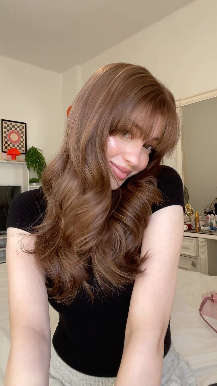 Woman showcasing long, wavy caramel-brown hair with curtain bangs indoors
