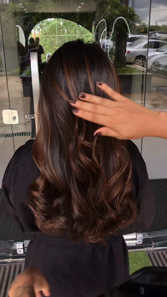 Long chocolate-brown curls show subtle caramel balayage highlights in a salon chair