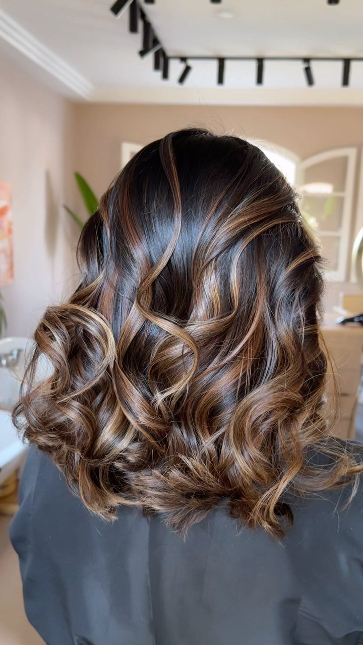 Dark brown curls with caramel highlights styled over a blue salon cape
