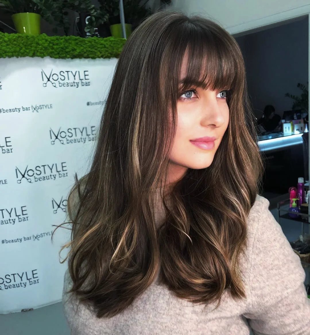 Woman with long wavy brown hair and straight blunt bangs in a salon