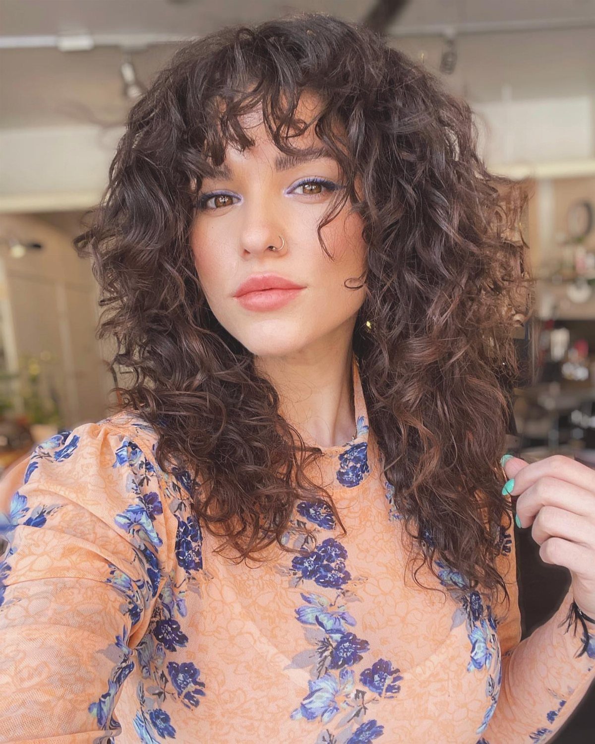 Woman with long layered dark curls and curly bangs wearing a peach floral top