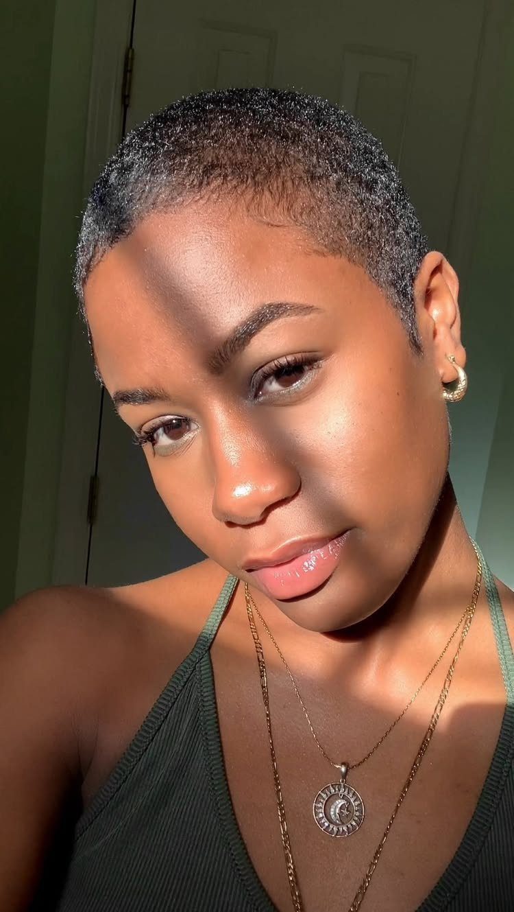 Person with very short black hair wearing gold earrings and layered necklaces