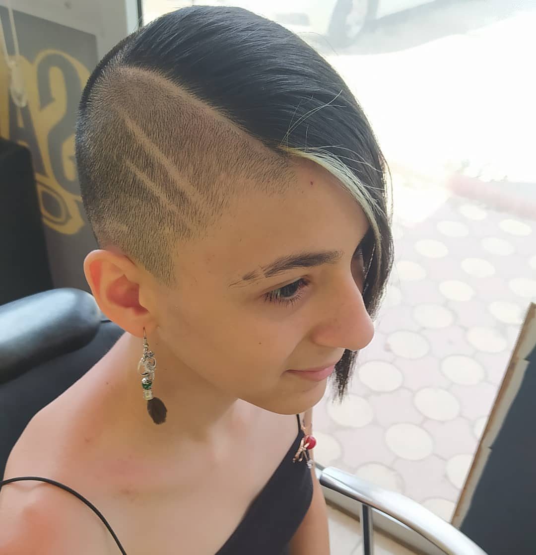 Long black side-swept hair contrasts with a shaved side featuring curved designs
