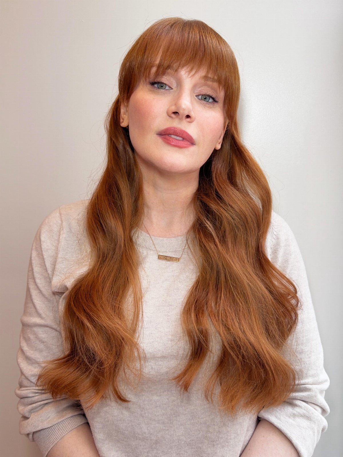Woman with long wavy reddish-brown hair and bangs against a light wall
