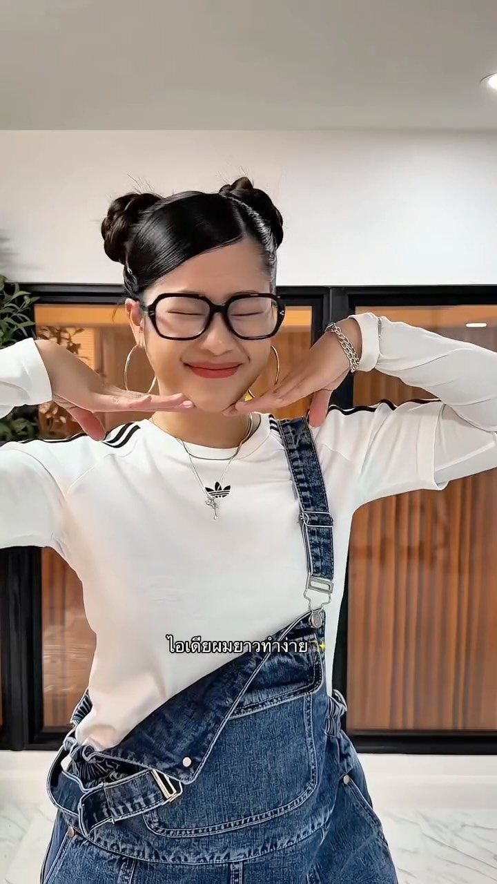 Woman wearing two high black hair buns, glasses, a white shirt, and denim overalls