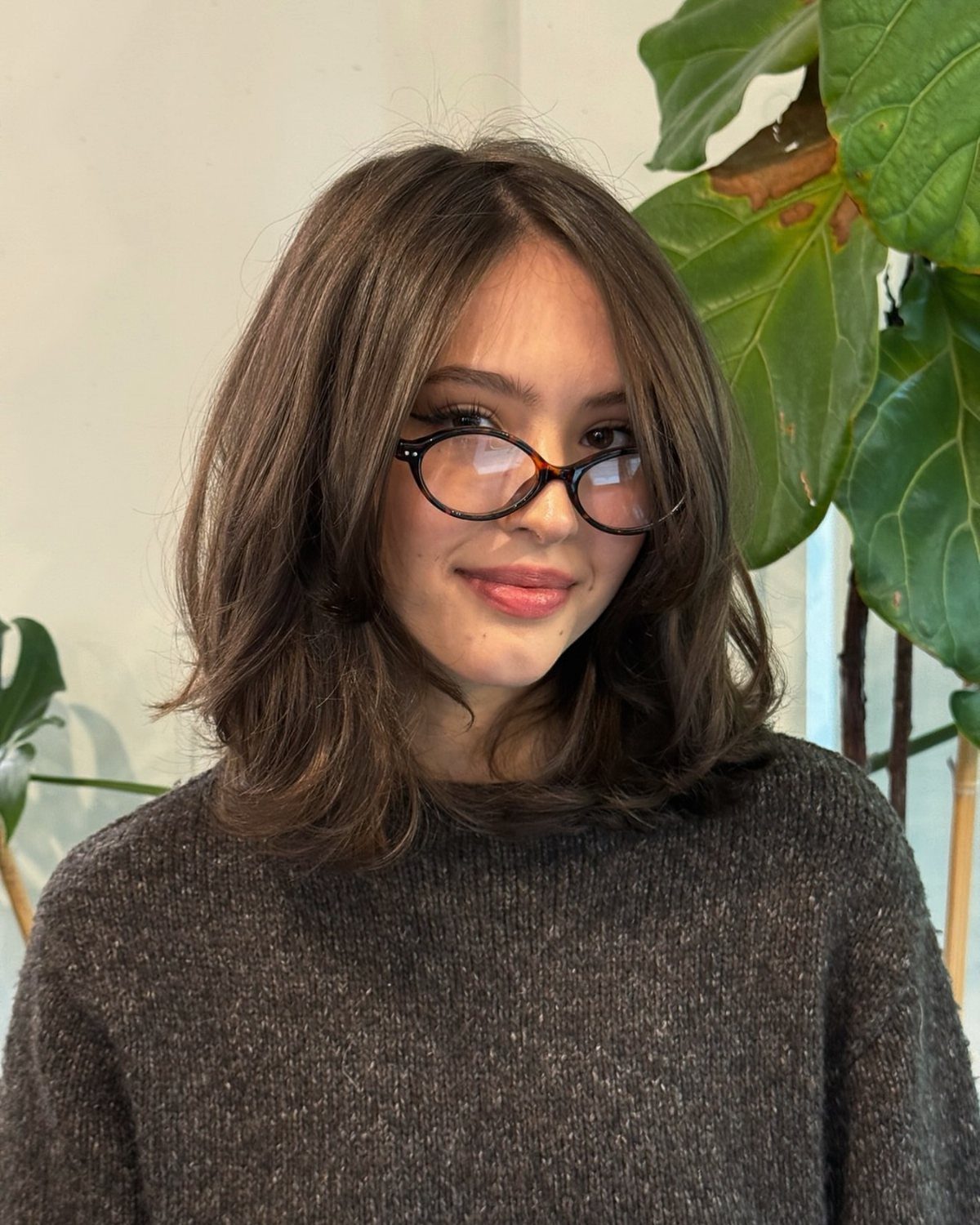 Person with layered shoulder-length brown hair and round glasses posing beside a leafy plant