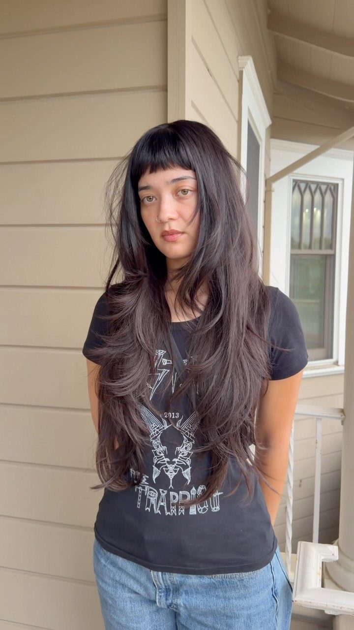 Person with long dark layered hair and blunt bangs standing on a porch