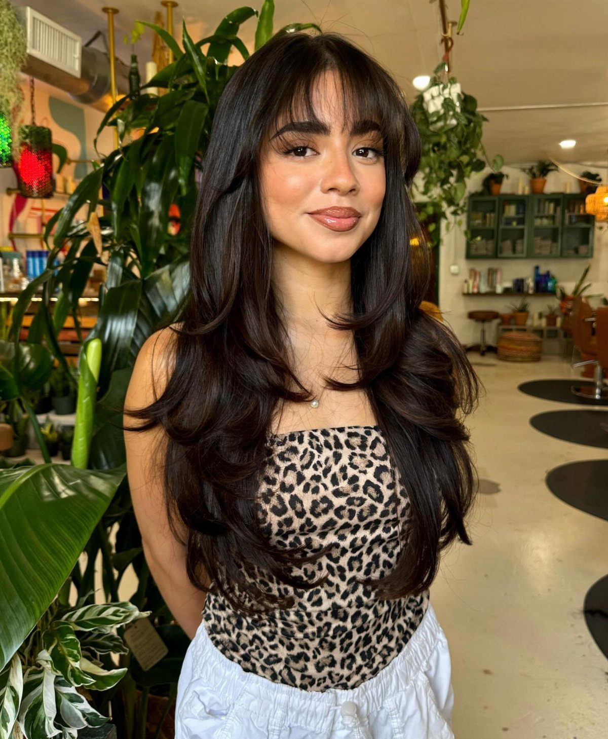 Woman with long dark layered hair and wispy blunt bangs posing among indoor plants