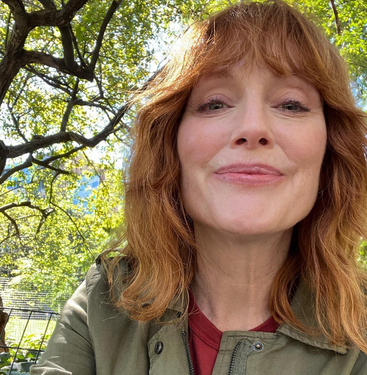 Woman with copper-red hair and bangs takes a selfie beneath leafy trees.