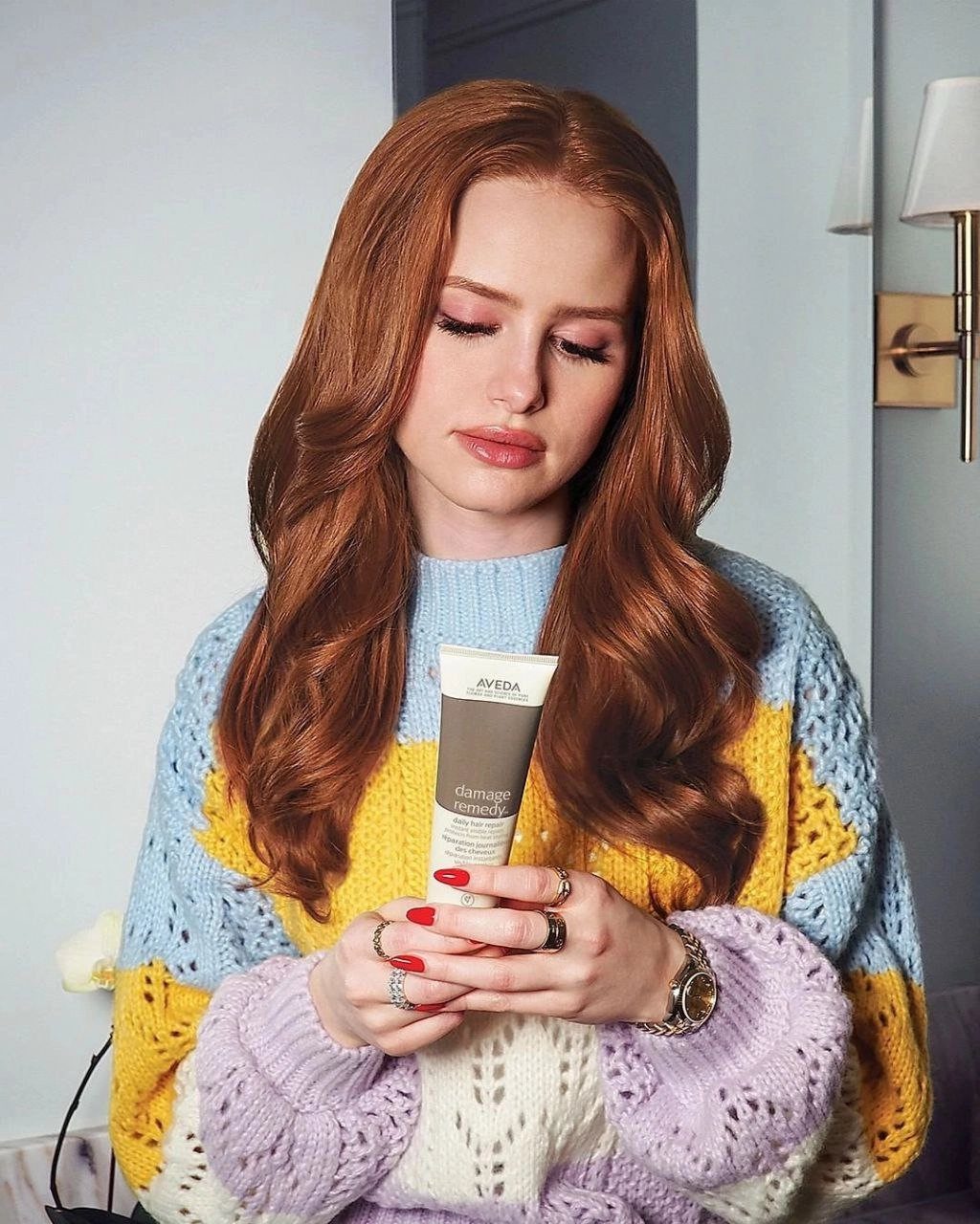 Woman with long copper-red hair holds a skincare tube in a colorful knit sweater