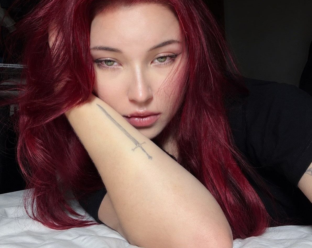 Woman with long, wavy violet-red hair resting her arm across her chest