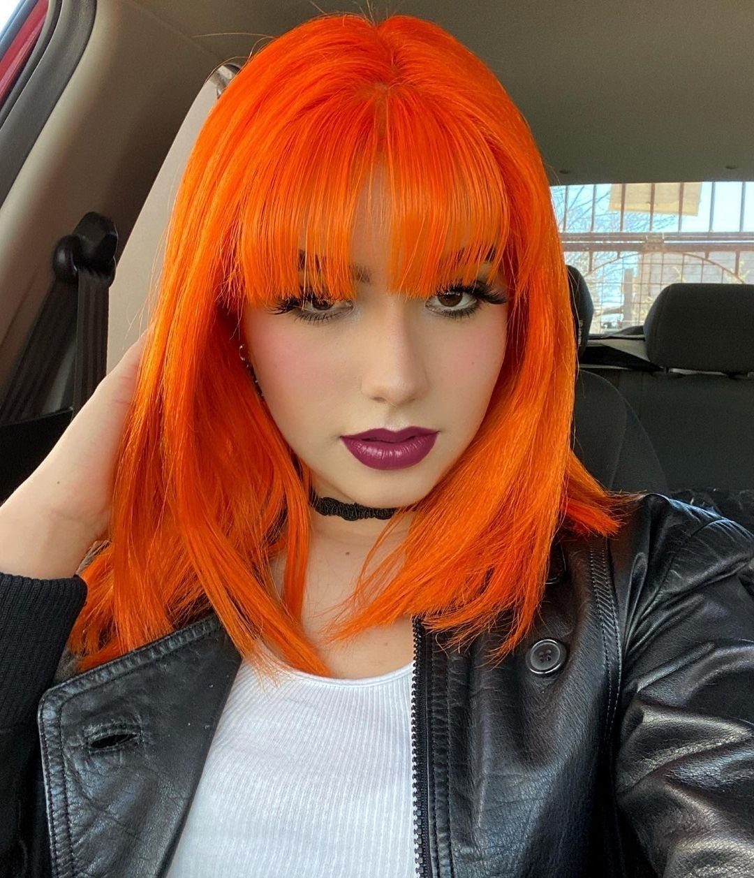 Vivid orange-red hair with blunt bangs frames a person posing in a car
