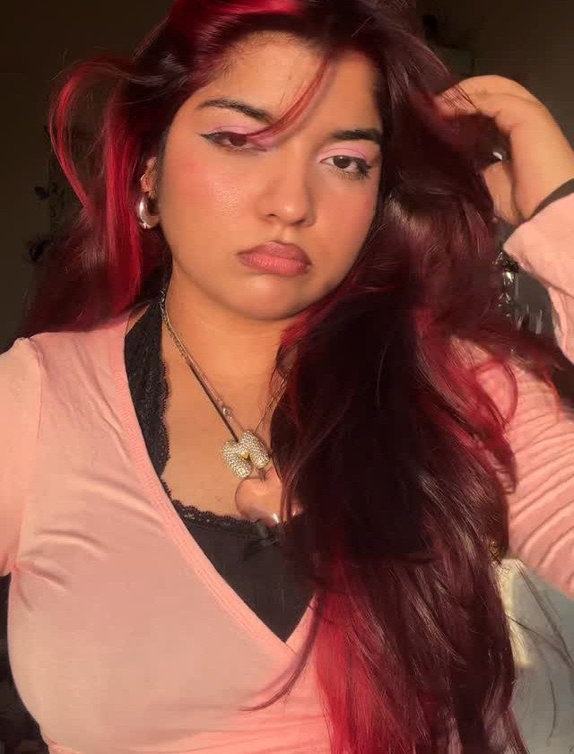 Long burgundy hair with vivid pink-red streaks frames a person wearing a pale pink top
