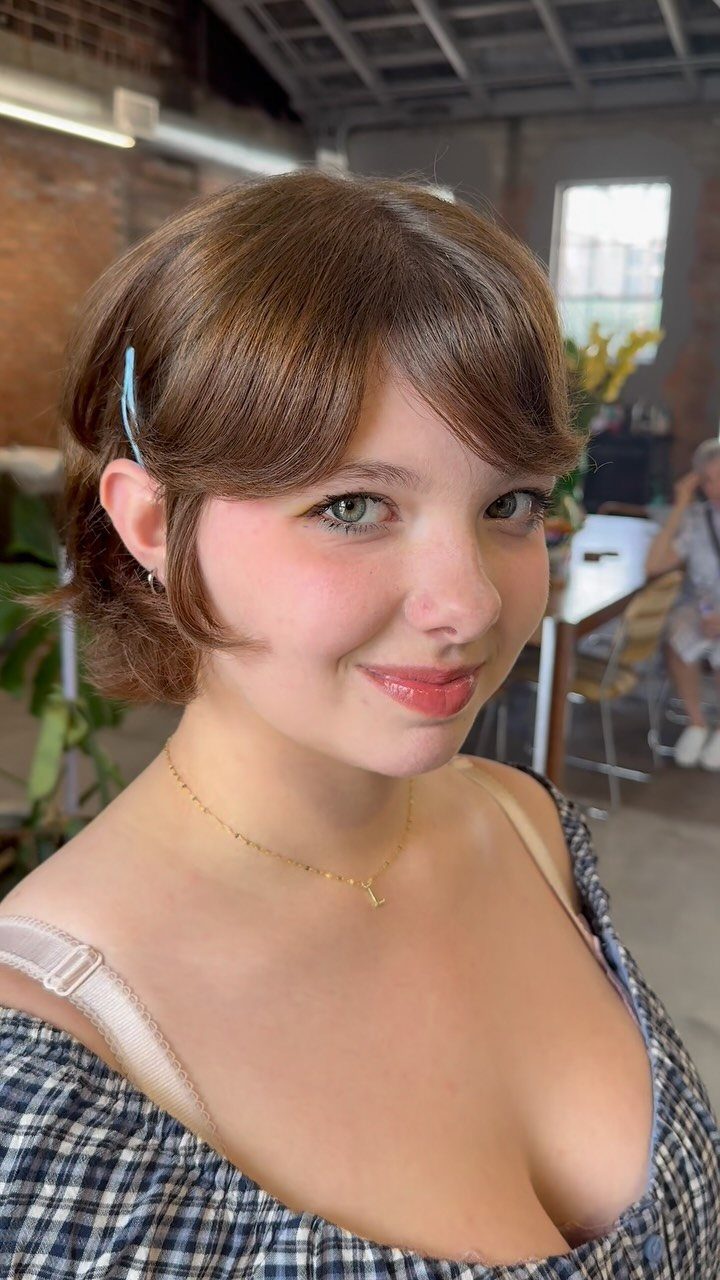 Young woman with short brown waves and a blue hair clip poses indoors
